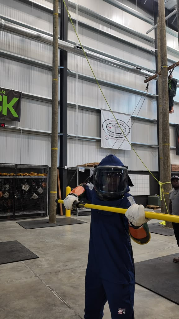 Trainee in a full 40 cal arc-flash suit and hood holding a yellow hot stick, with a climber on a pole behind
