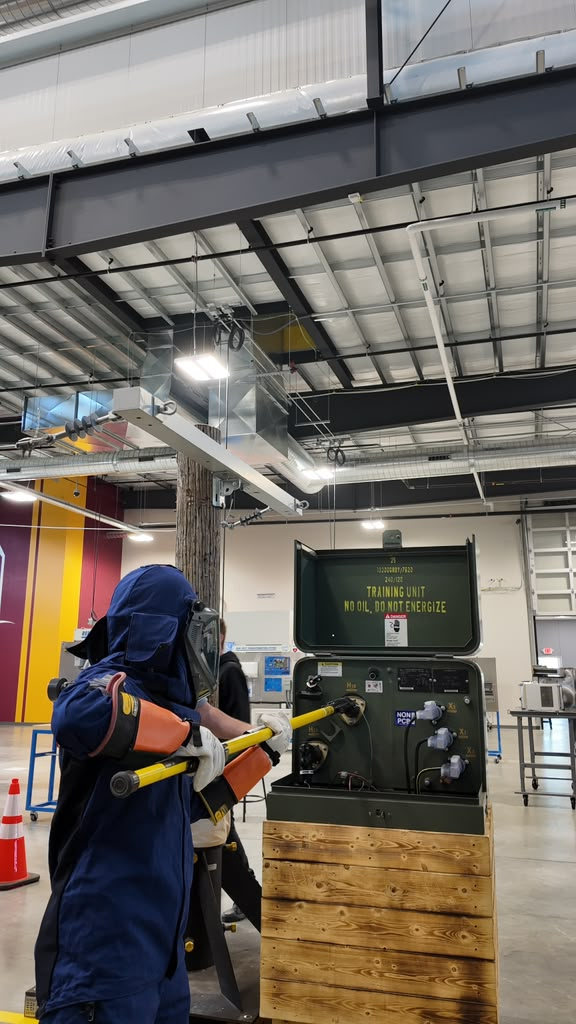 Trainee in arc-rated PPE operating a hot stick at a pad-mounted transformer under instruction