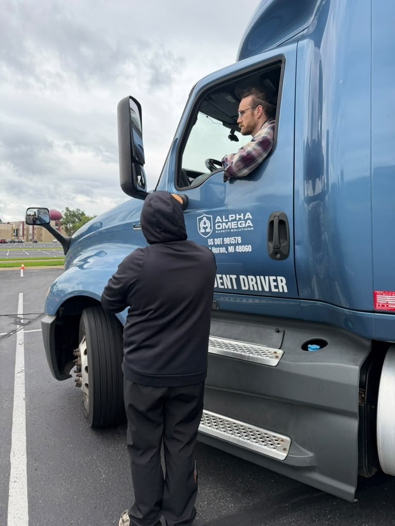 CDL instructor seated in the cab of the blue student-driver tractor talking down to a student standing at the door