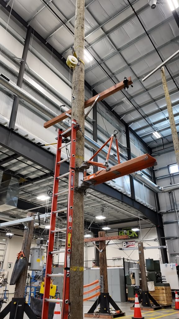 Indoor training pole with an extension ladder, rigged crossarm and lifting gear, transformers and cones behind