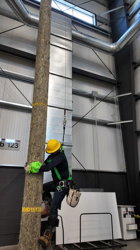 Trainee in a green safety harness climbing a pole inside the indoor training facility