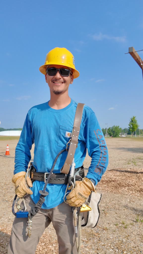 Lineworker student in a yellow hard hat, sunglasses and full climbing harness standing in the training yard