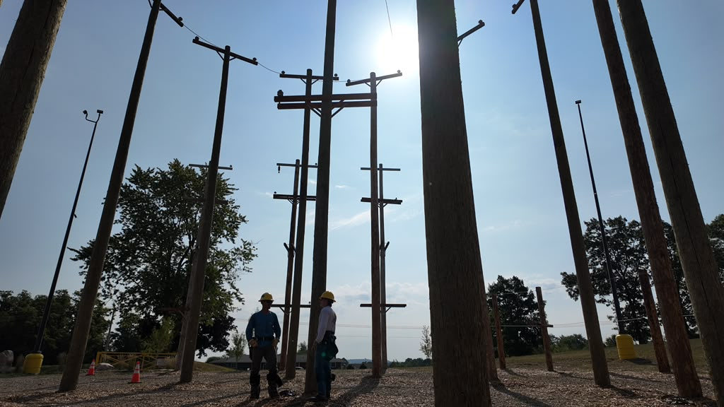 Two crew members in hard hats standing among the wooden poles of the outdoor training yard, backlit by a low sun
