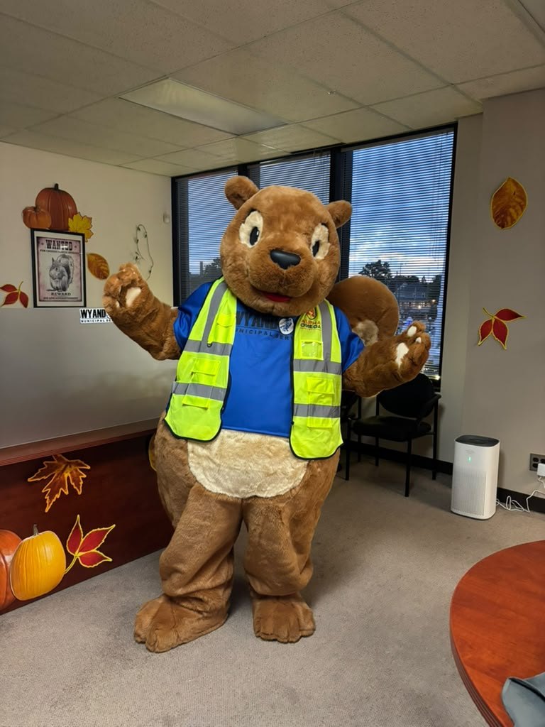 A squirrel mascot in a blue Wyandotte Municipal Services shirt and a yellow hi-vis vest, waving in an office decorated for autumn